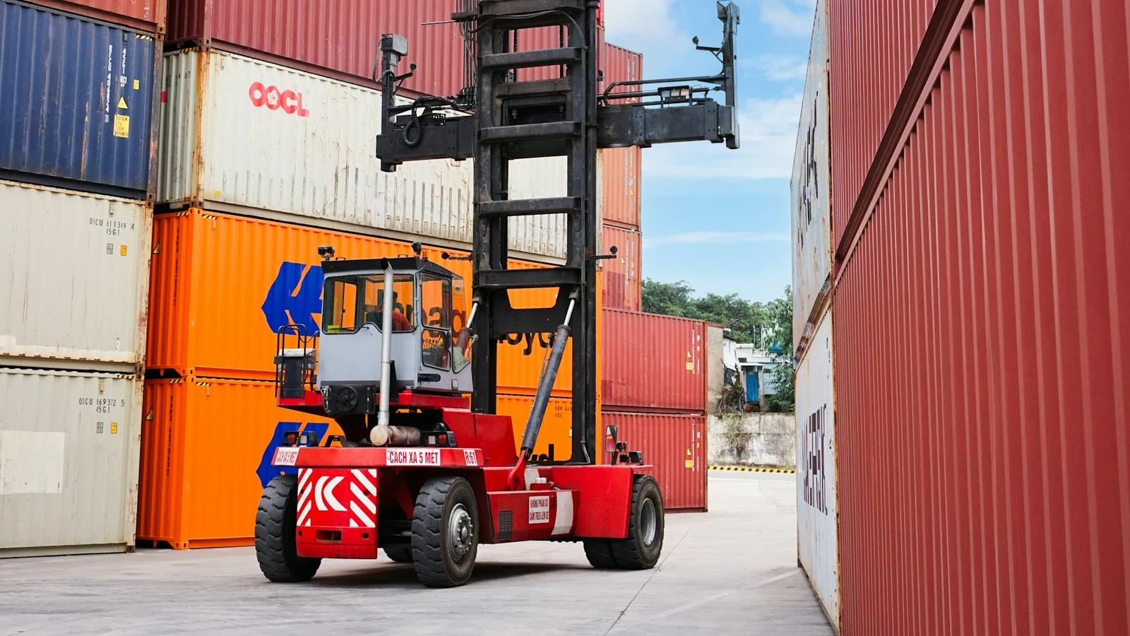 A red heavy-duty container handler with a raised mast and spreader in a container yard, surrounded by stacked ocean shipping containers.
