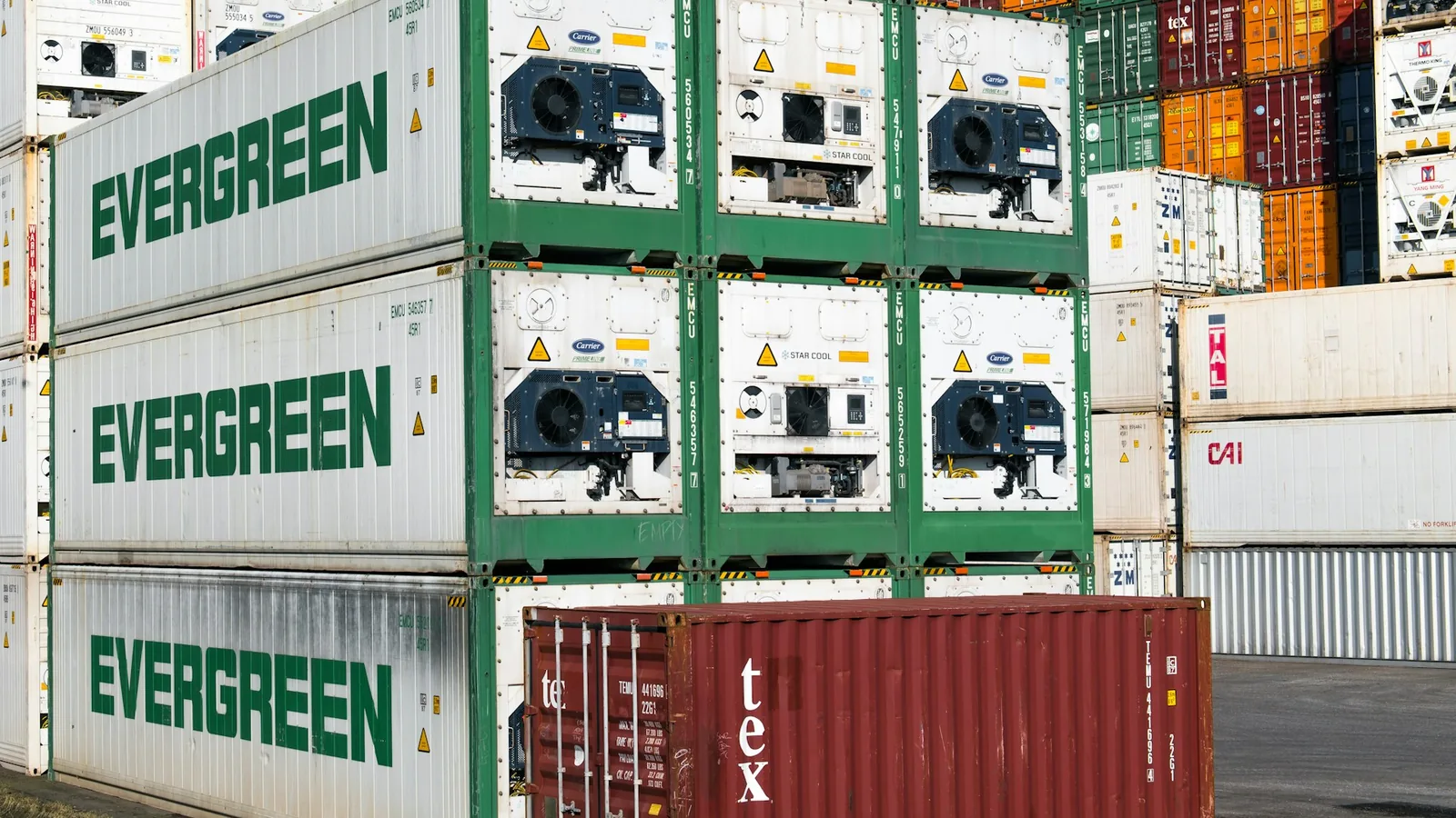 Stacked white refrigerated ocean shipping containers with their reefer refrigeration units visible at a marine container terminal.