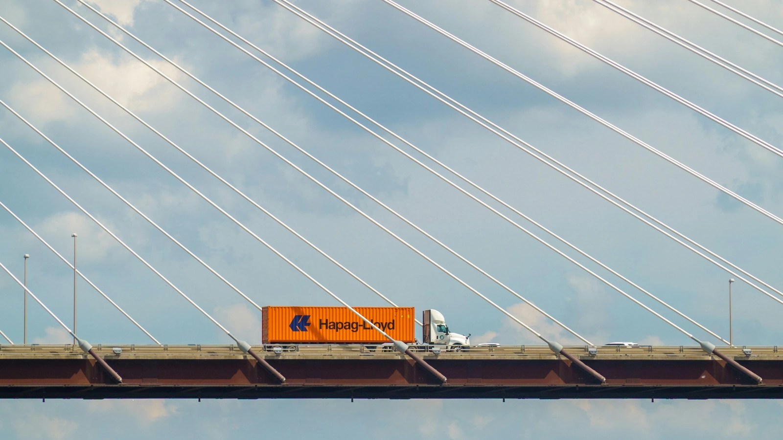 A white conventional sleeper-cab truck hauling an orange Hapag-Lloyd ocean shipping container across a cable-stayed highway bridge.
